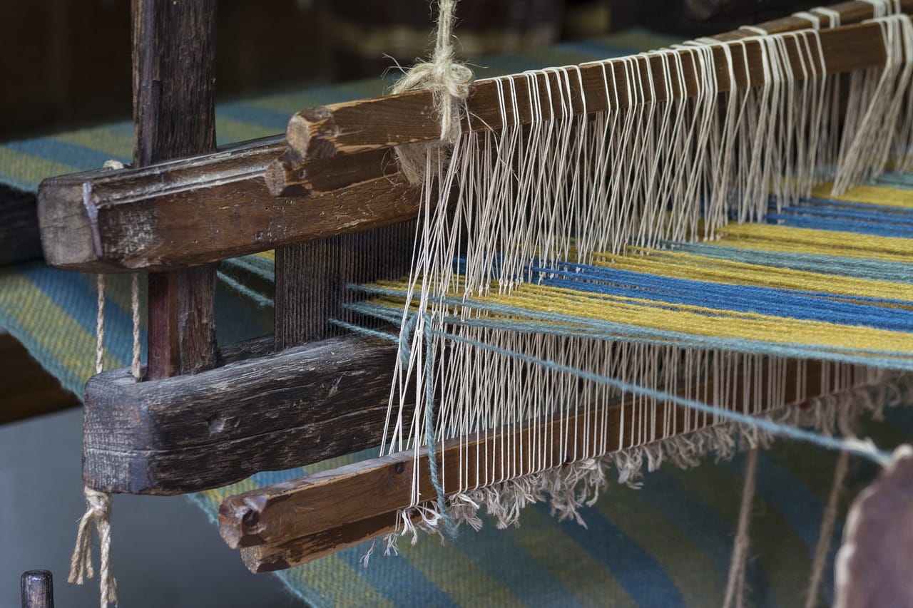 A close-up view of a traditional wooden loom with threads stretched taut across it. Yellow and blue yarns are interwoven with white warp threads, creating a striped textile pattern in progress.